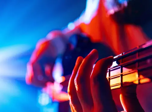 Close-up of a person playing an electric guitar, with fingers pressing the strings on the fretboard. The background features vibrant blue and orange stage lighting.