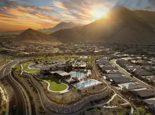 Aerial view of a residential neighborhood at sunset, surrounded by mountains. The community features a clubhouse, green lawns, and a swimming pool with lounge chairs. The sun is setting behind the hills, casting a warm glow.