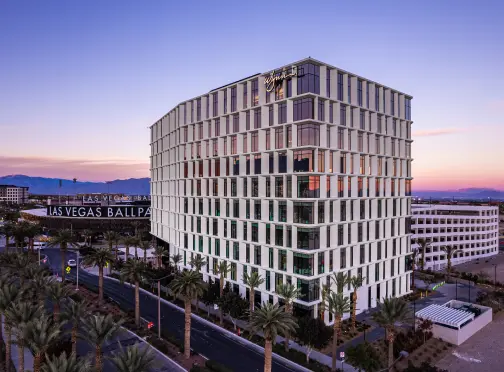 A modern, multi-story hotel building with a grid-like facade stands at sunset in Las Vegas, with palm trees lining the street and mountains visible in the background. A sign reads Las Vegas Ballpark.
