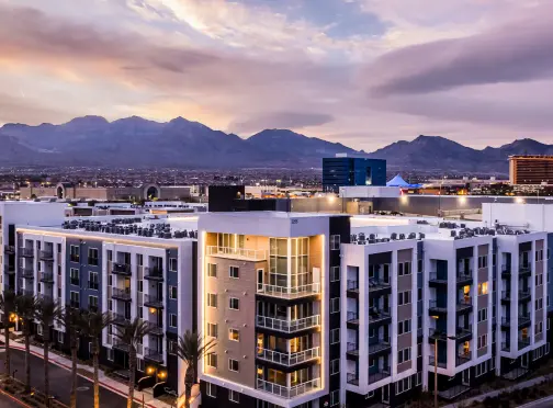 Modern mid-rise apartment buildings with lit balconies at dusk, palm trees in front, and mountains in the background under a colorful, cloudy sky.