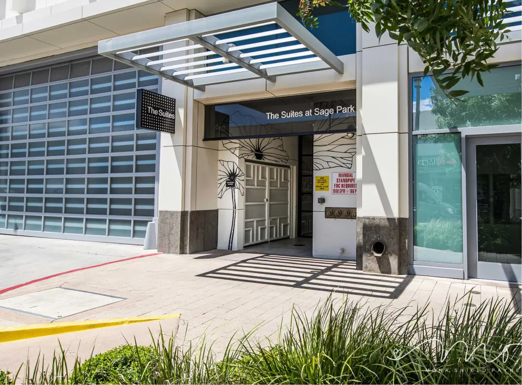 The entrance to The Suites at Sage Park, featuring a modern awning, wall art of flowers, and a sign above the door. The area is clean with greenery and a sidewalk in front.