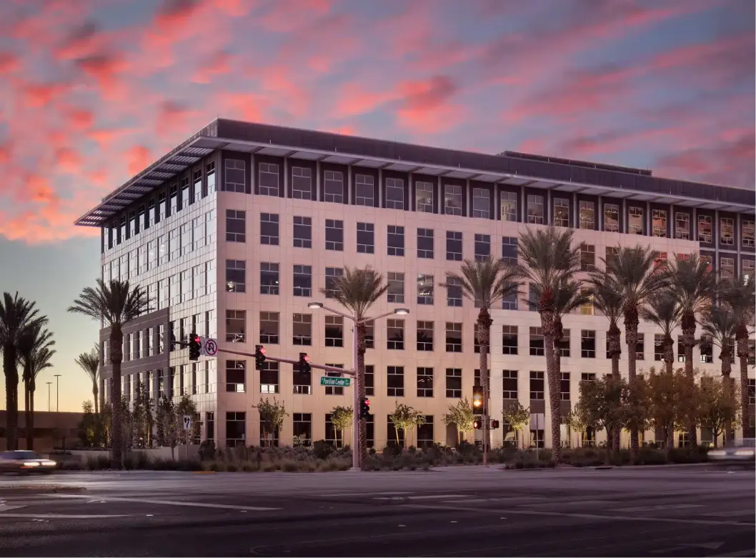 A modern, multi-story office building with large windows stands on a street corner lined with tall palm trees under a colorful sunset sky with pink clouds. Traffic lights and street signs are visible in the foreground.