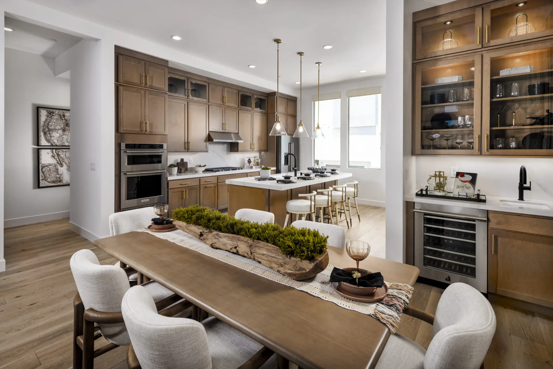 Modern kitchen and dining area with wooden cabinets, built-in appliances, pendant lighting, a long dining table with white chairs, centerpiece greenery, and a wet bar with glass-front cabinets and a wine cooler.