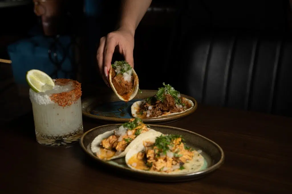 A hand lifts a taco from a plate on a dark table with three tacos garnished with cilantro and onion. Nearby, there is a margarita cocktail with a chili-lime rim and a lime wedge.