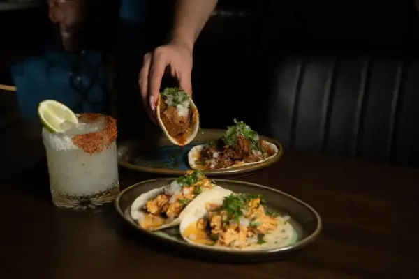 A hand picks up a taco filled with meat and cilantro from a plate. Two more tacos and a glass of a margarita garnished with lime and chili salt sit on a dark wooden table.