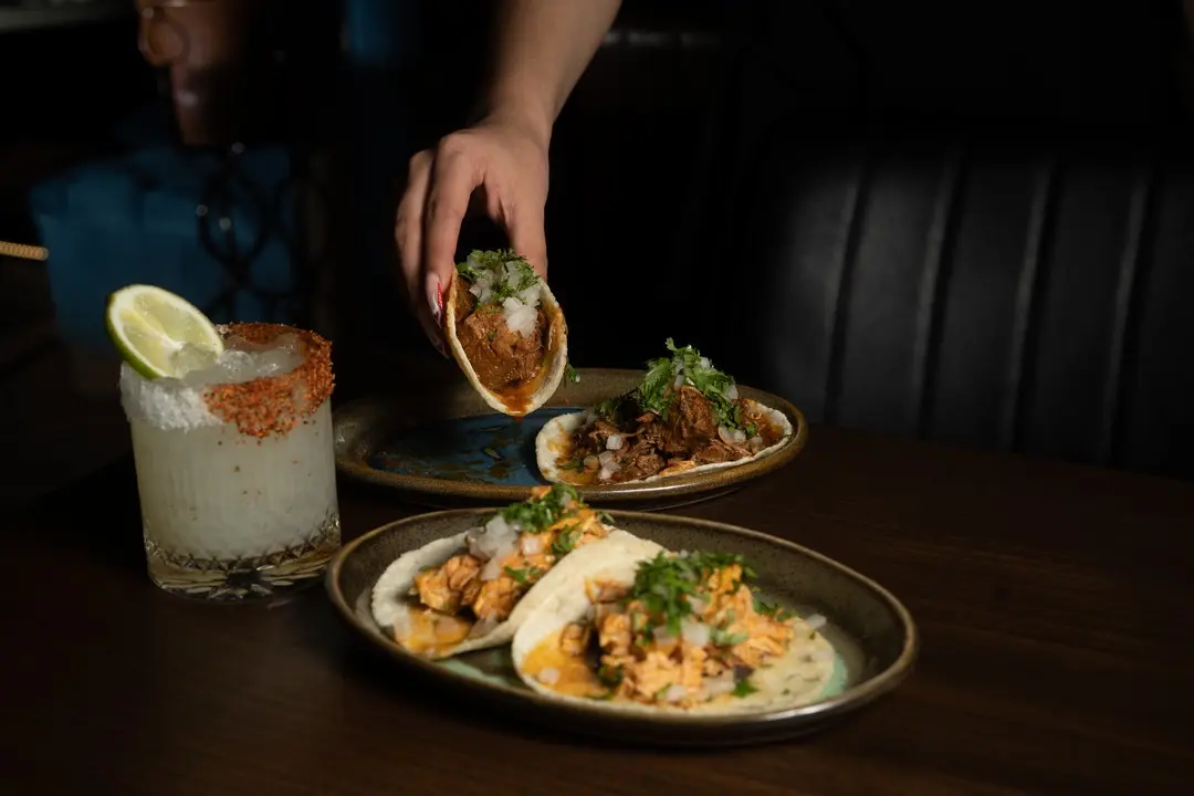 A hand picks up a taco filled with meat and cilantro from a plate. Two more tacos and a glass of a margarita garnished with lime and chili salt sit on a dark wooden table.