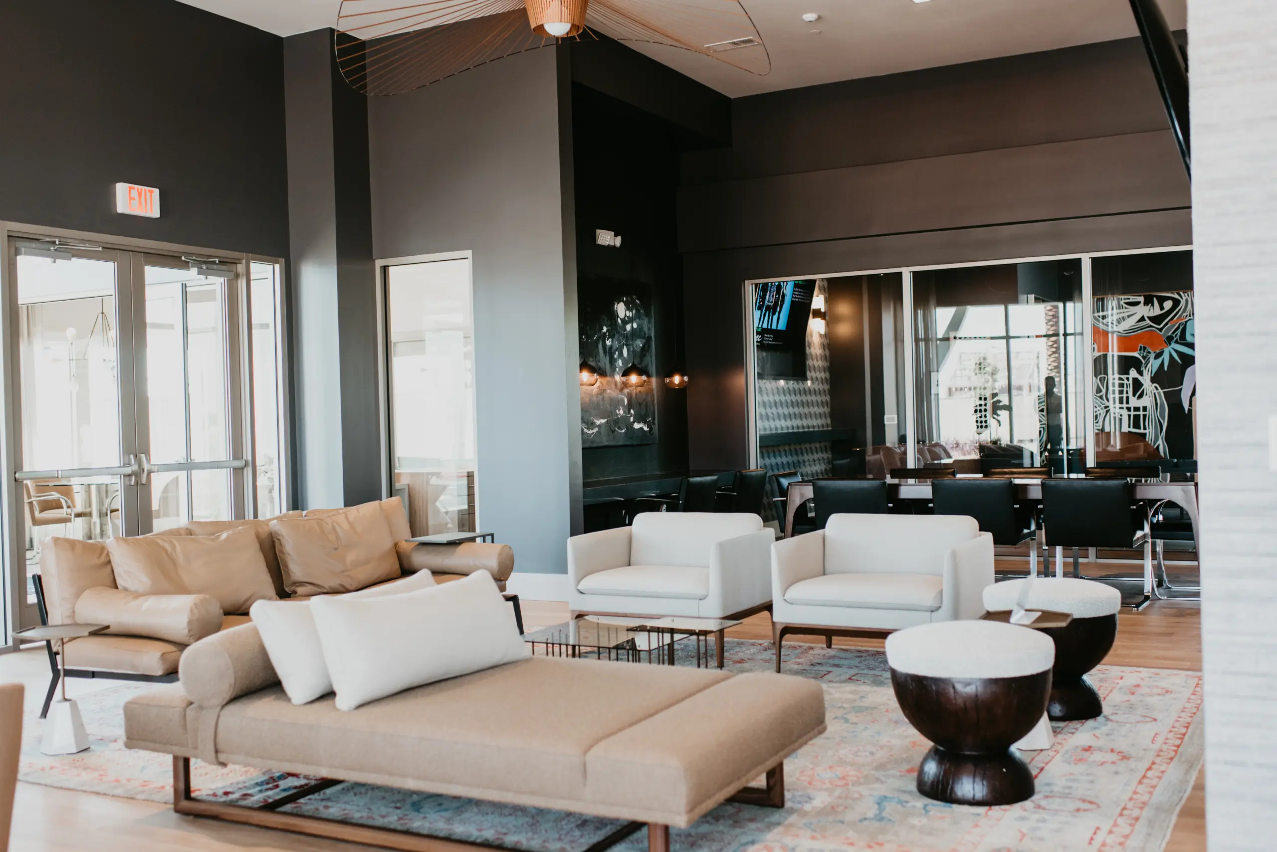 1d1b0ffd97a56bb04d78bb60c42c41876c51dfd8 A modern lounge area with beige sofas, white armchairs, small round tables, and a contemporary rug. Glass doors and windows let in natural light, and a conference room is visible in the background.