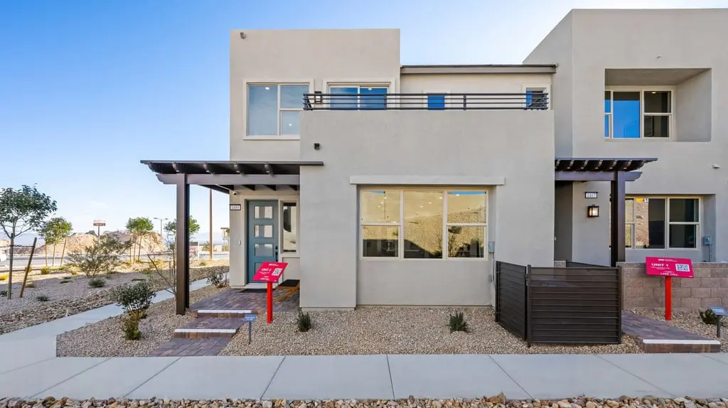 Modern two-story townhouse with light-colored stucco exterior, large windows, a small covered porch, and minimal desert landscaping featuring gravel, rocks, and a few plants. Two red real estate signs are visible in front.