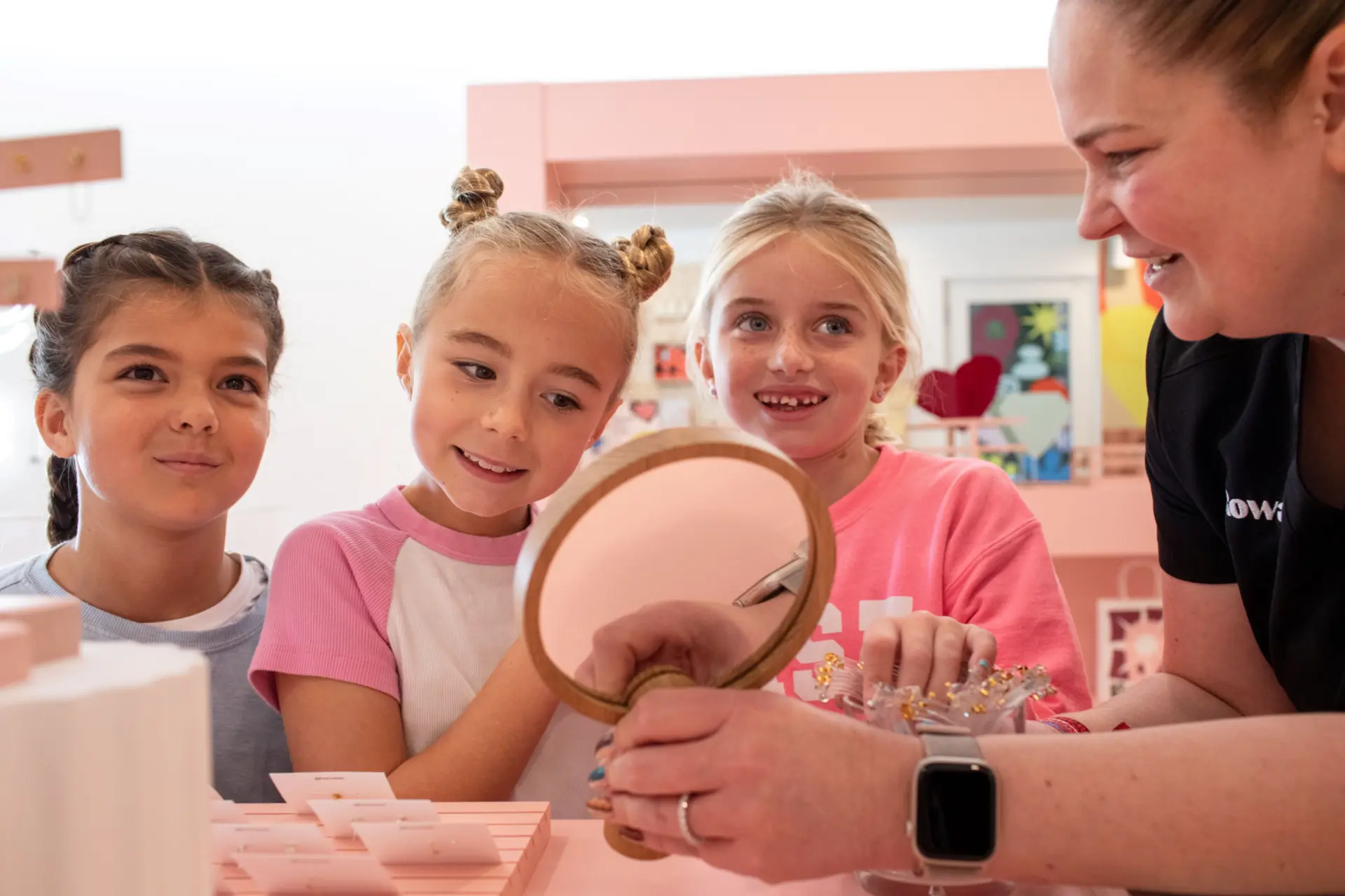 Three young girls smile and look at a mirror held by an adult woman, who is helping them in a brightly lit, pink-themed room, possibly a jewelry or craft store. The atmosphere is cheerful and engaging.