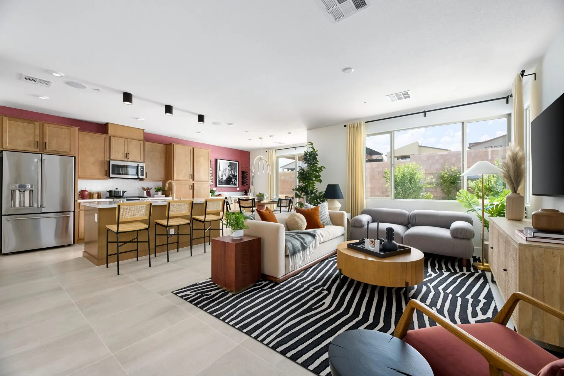 Modern open-concept living room and kitchen with wood cabinets, stainless steel appliances, bar stools, a cozy seating area, large windows, and abundant natural light. Black-and-white rug adds contrast to the neutral decor.