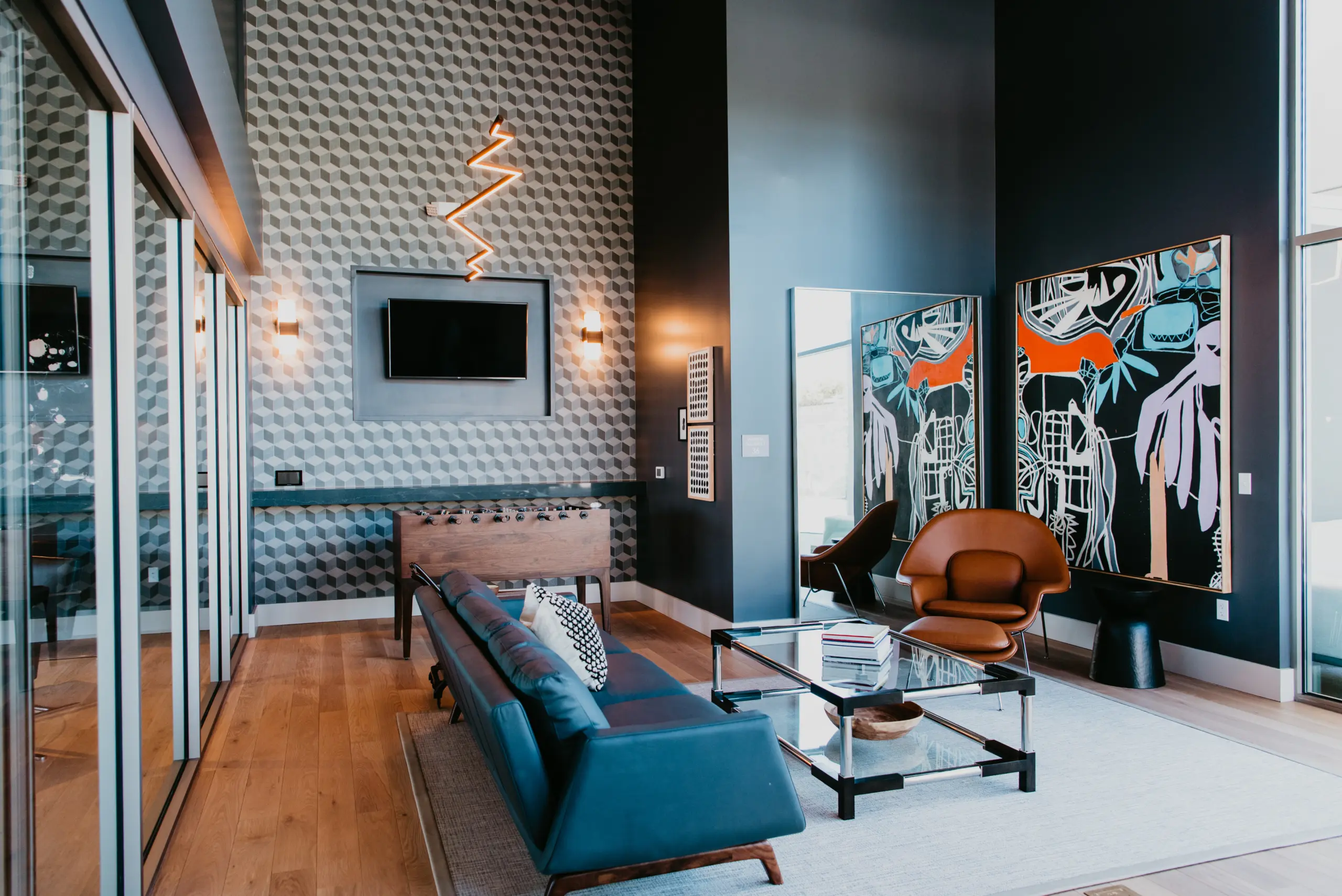 2d0c4a9a82be0e55c85ac7f9057cdb0880c5425b A modern living room with geometric patterned wall, abstract artwork, blue sofa, glass coffee table, foosball table, and wood flooring. Warm lighting and large windows create a bright, stylish space.