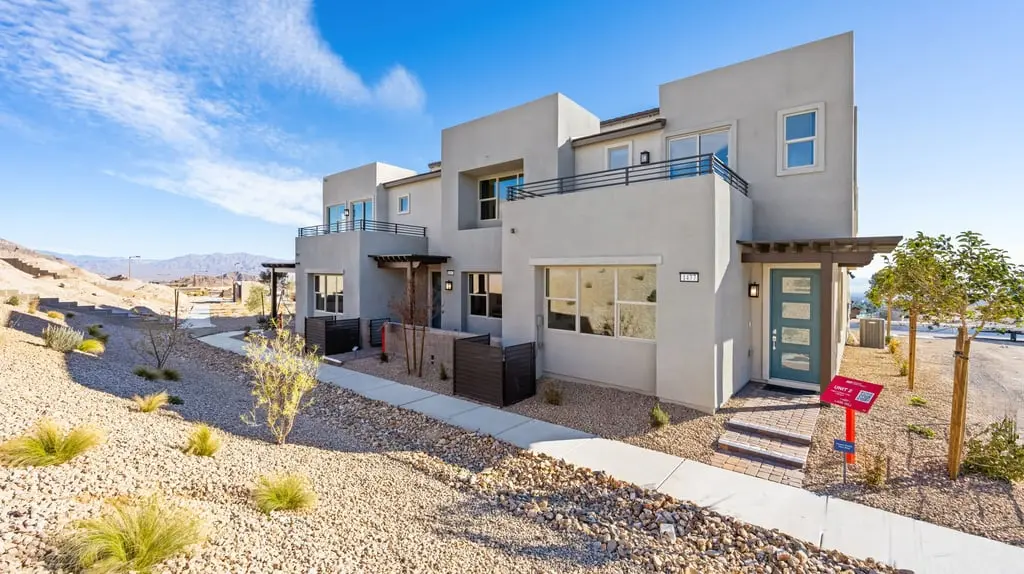 Modern two-story house with a flat roof, large windows, and a balcony. Desert landscaping with rocks and sparse plants surrounds the exterior. Bright blue sky in the background.