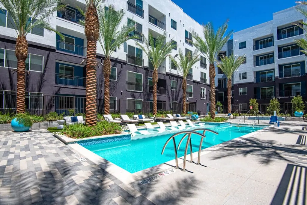 3edfde52311b2224312a8be4145b53d91573fcb1 Modern apartment complex with tall palm trees surrounding a rectangular swimming pool, featuring in-water lounge chairs, poolside seating, and a tiled deck under a clear blue sky.