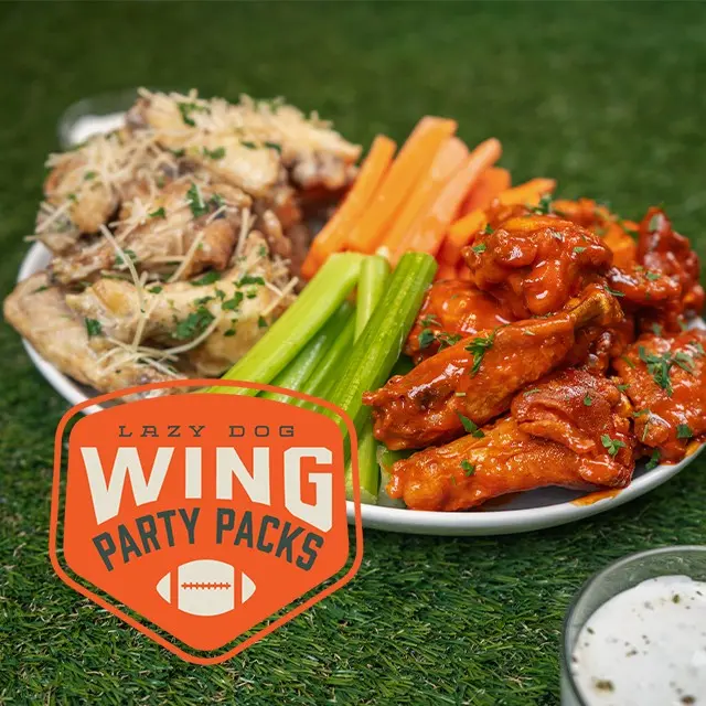 A plate of chicken wings with two flavors, celery, and carrot sticks sits on green turf. The text Lazy Dog Wing Party Packs with a football icon is overlaid. A small cup of ranch dressing is in the foreground.