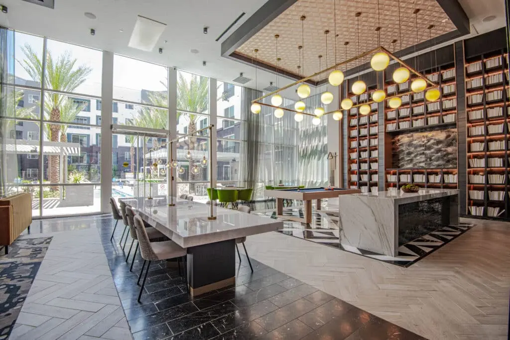 6f808aa16dd130a6d16e99220a211ebaea214432 Modern, stylish lobby with marble tables, plush chairs, a geometric chandelier, large bookshelf wall, and floor-to-ceiling windows overlooking a courtyard with palm trees and a pool.
