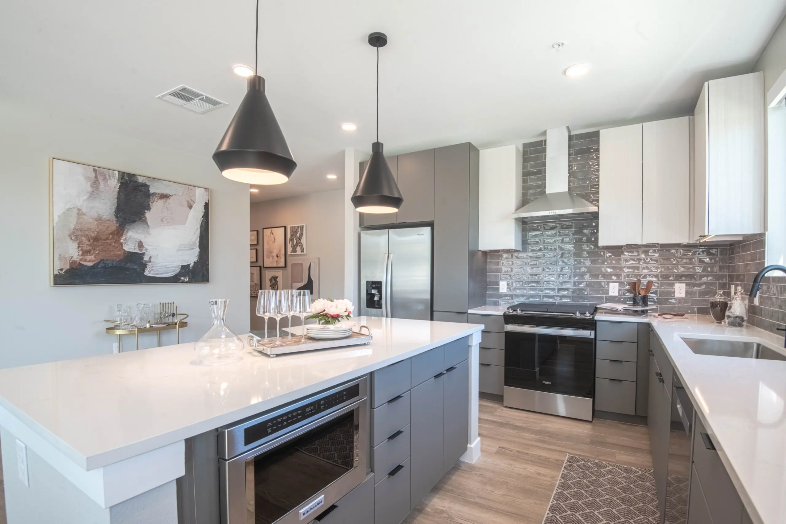 7ec6b972a06fcb3a3caefa2880cb7897f925d49e Modern kitchen with gray cabinets, white countertops, stainless steel appliances, pendant lights over an island with barware, a built-in microwave, and neutral decor, including abstract art on the wall.