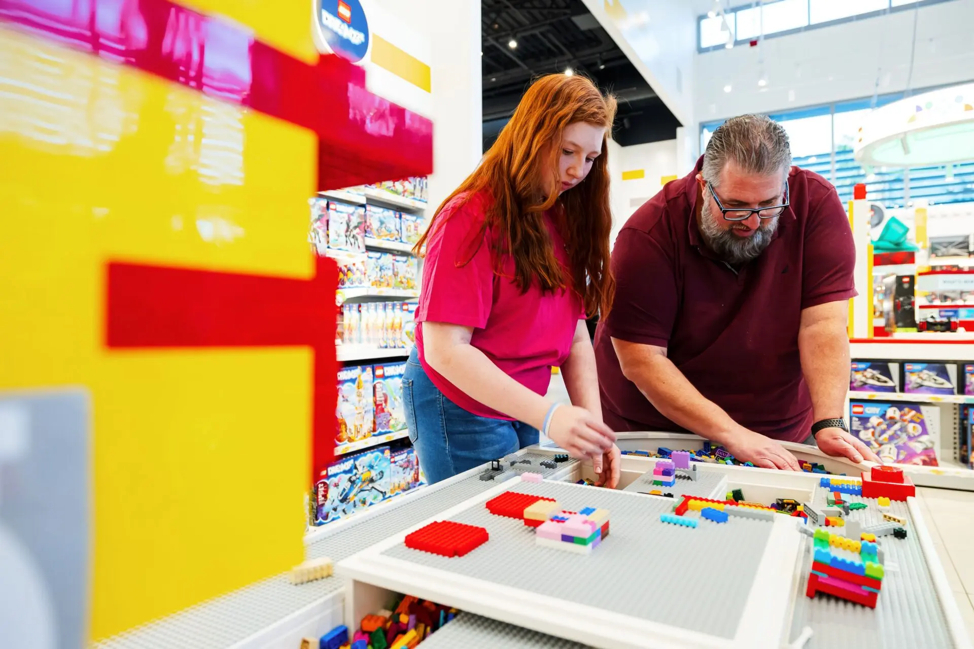 Arthur Rossi Lego master A young woman and an older man build with colorful LEGO bricks at a play table inside a LEGO store, surrounded by LEGO sets on display shelves.