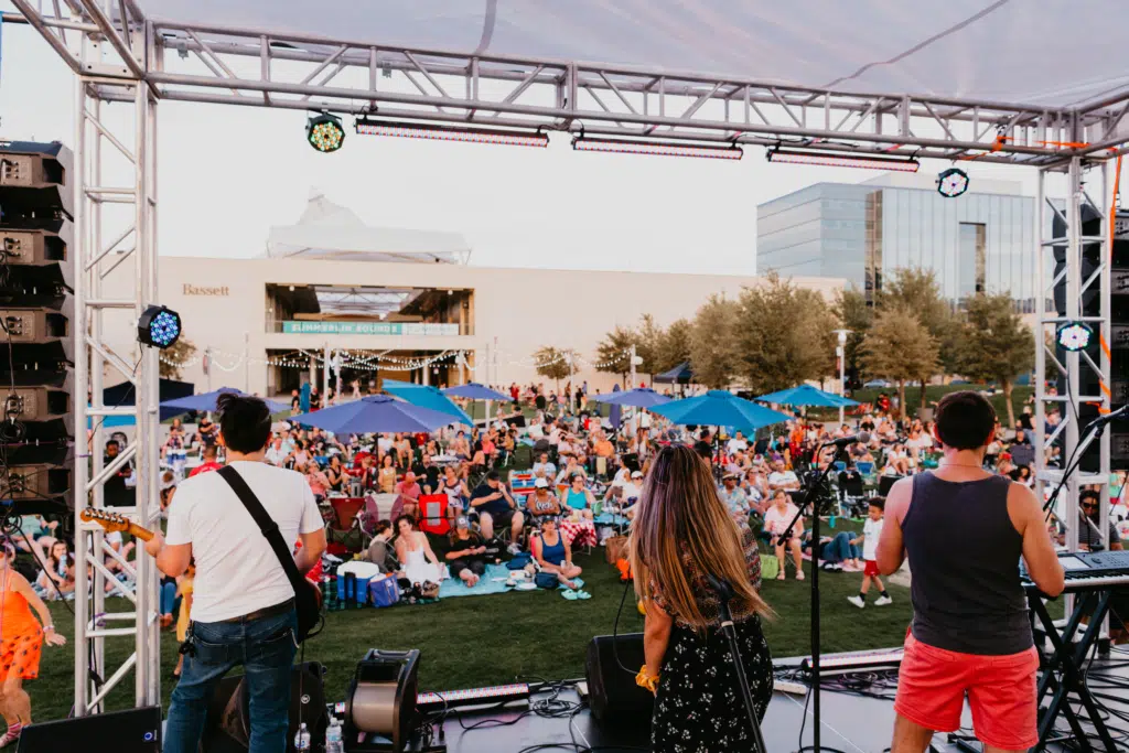 View from behind a band performing on an outdoor stage, looking out at a large crowd sitting and standing on a grassy lawn, with blue umbrellas and modern buildings in the background.