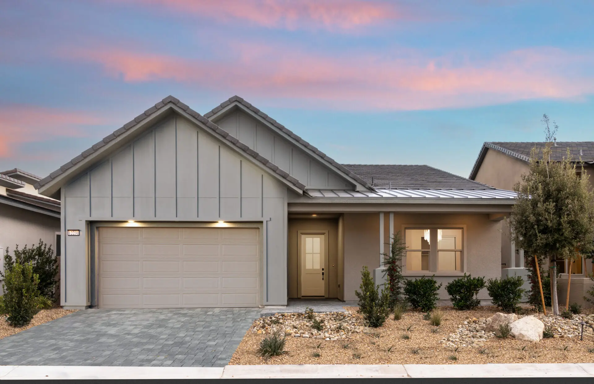 Modern single-story house with light gray siding, double garage, and a small front porch. The yard features desert landscaping with rocks and drought-tolerant plants, under a colorful sunset sky.