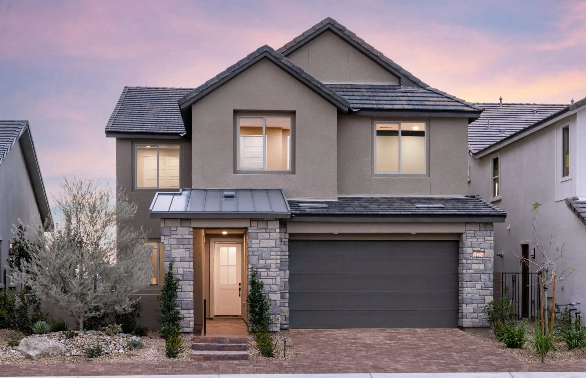 Two-story modern house with gray siding, stone accents, a dark gray garage door, and large windows. The home is surrounded by shrubs and a small tree, with a paved driveway at sunset.