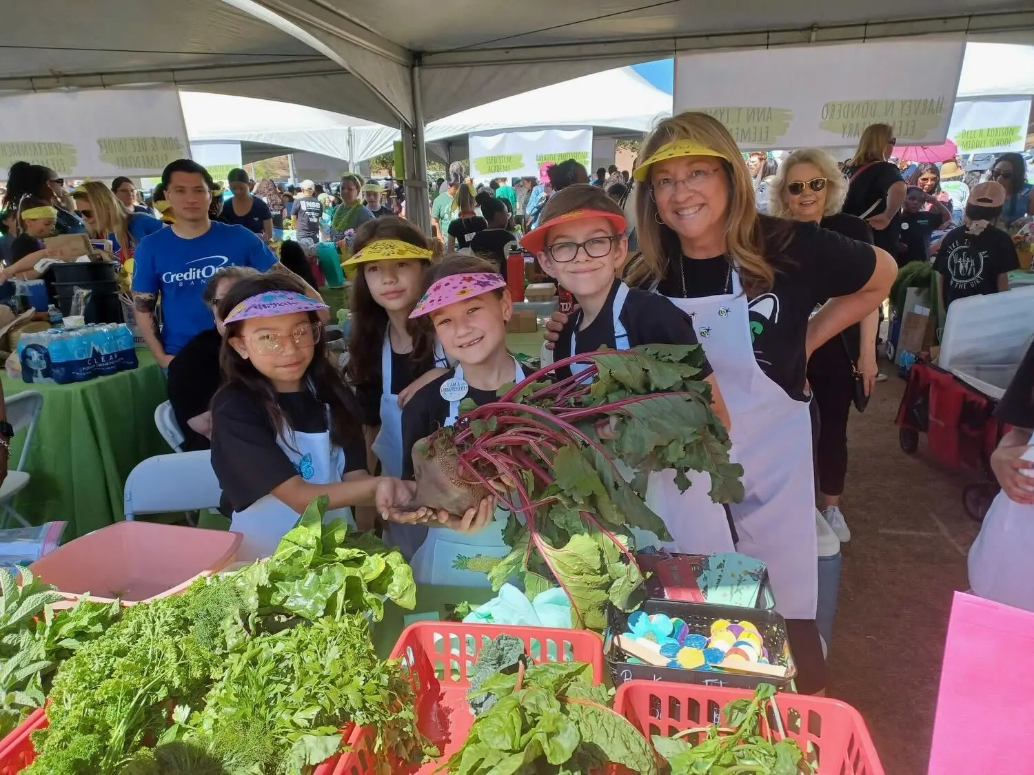 A group of children and an adult, all wearing aprons and visors, smile while holding a large beet in a tented outdoor event. Baskets of fresh greens and produce are on the table in front of them.