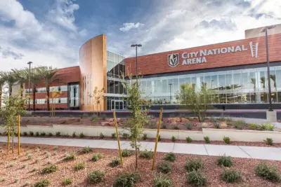 City National Arena building with modern glass and metal facade, landscaped with small plants and palm trees, under a partly cloudy sky.