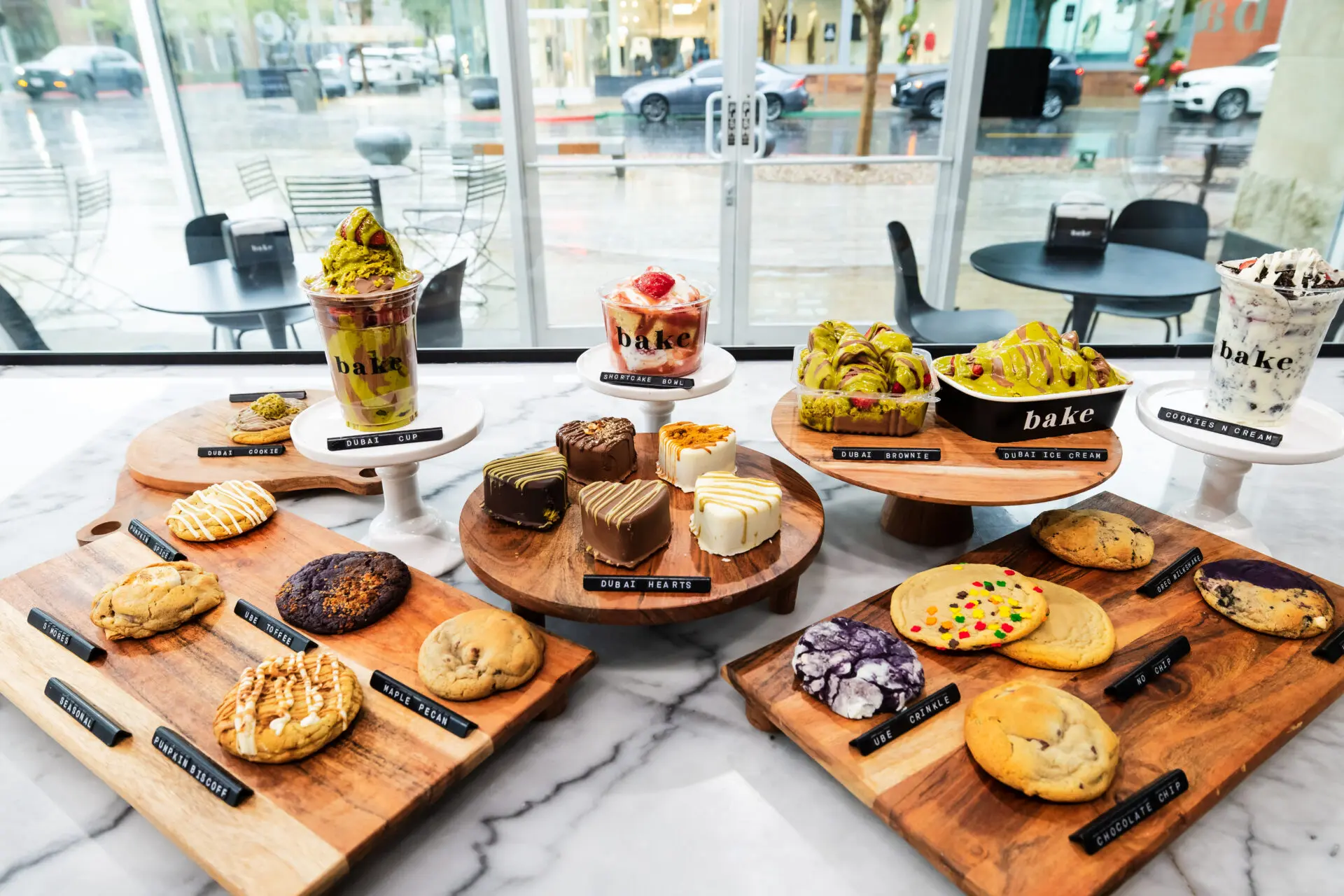 A variety of cookies, brownies, and ice cream desserts are displayed on wooden boards atop a marble counter in a bright, modern bakery with large windows and outdoor seating visible.