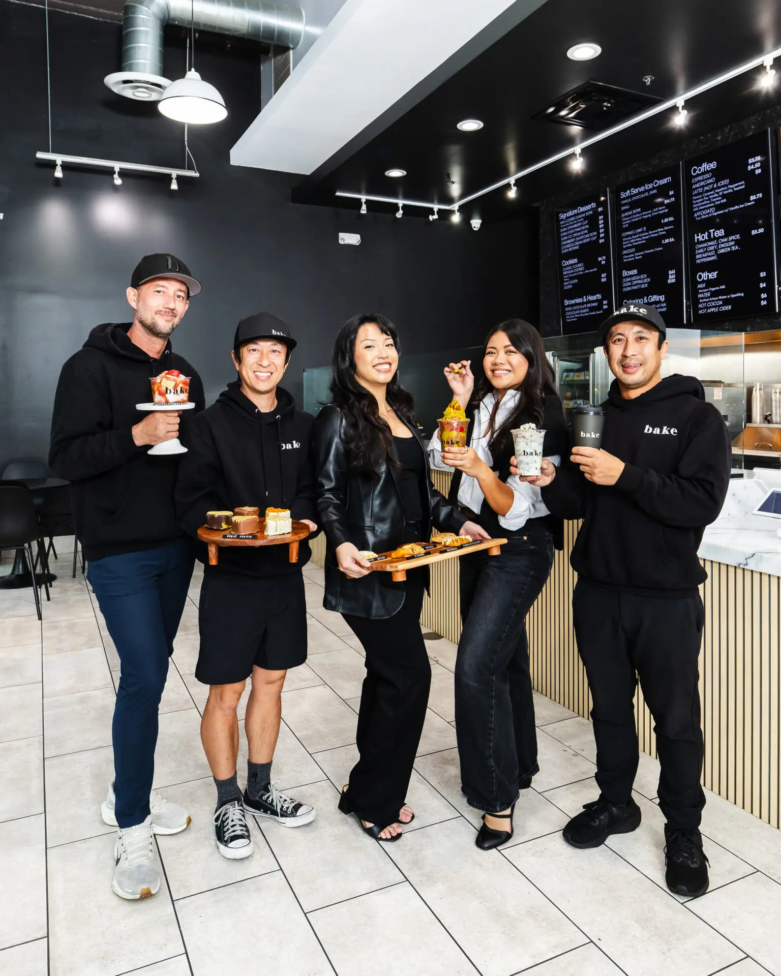 Five people stand in a modern dessert shop, smiling and holding various desserts and drinks. The shop has a black and white interior with a menu on the wall and a counter with display items.