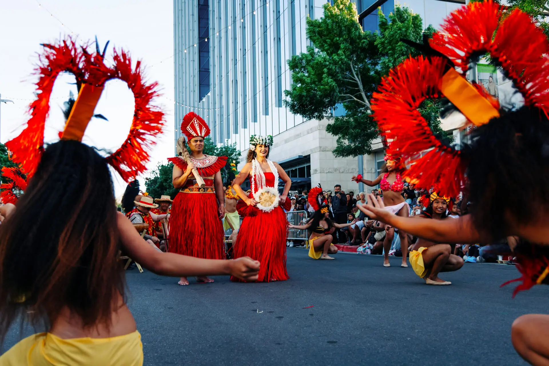 Dancers in vibrant red and yellow costumes perform a traditional Polynesian dance in the street, with onlookers in the background and tall buildings and trees lining the scene.