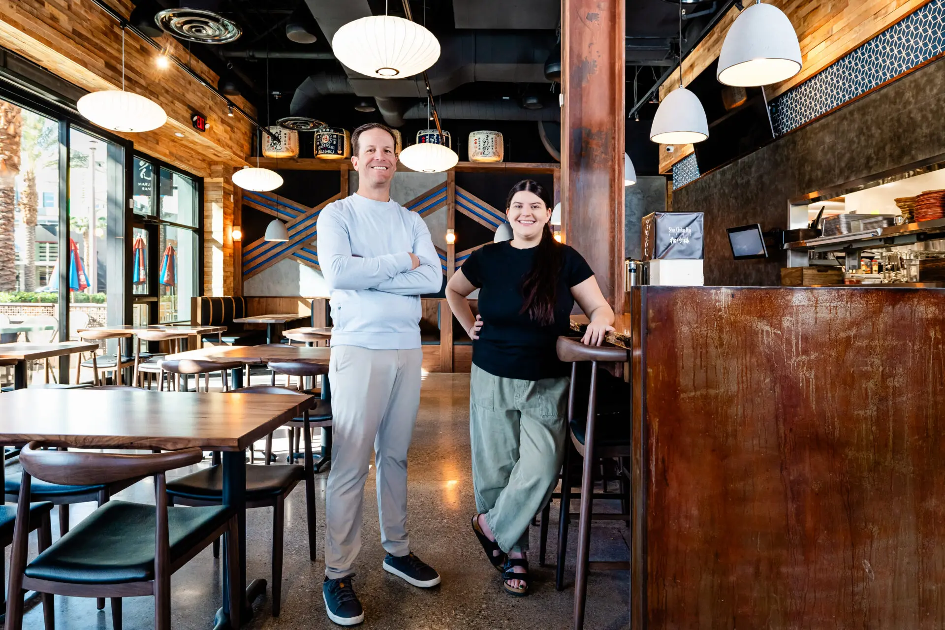 Two people, a man and a woman, stand smiling inside a modern, stylish restaurant with wooden tables and hanging lights. The restaurant appears bright and welcoming with large windows and contemporary decor.