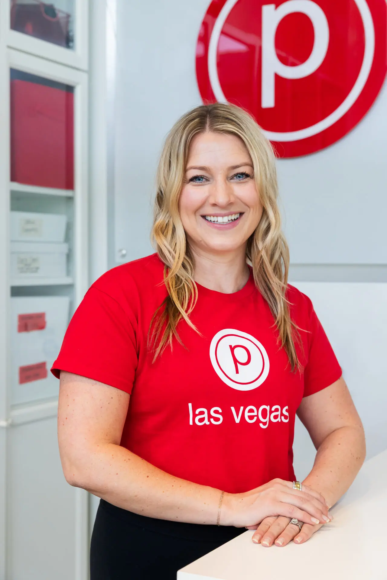 A smiling woman with long blonde hair stands at a counter, wearing a red las vegas t-shirt with a circular p logo. A matching logo sign is on the wall behind her. The setting appears to be a modern store or reception area.