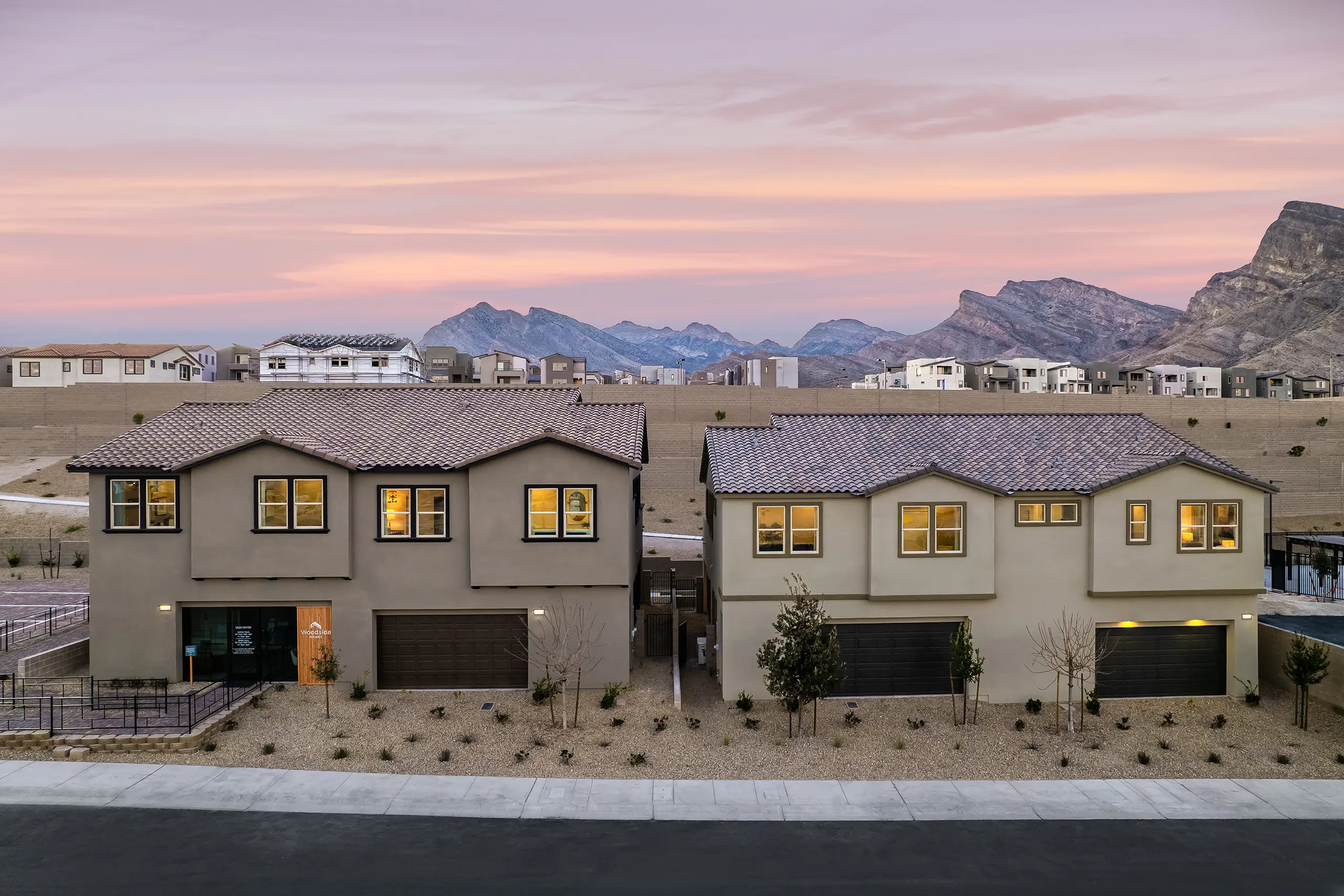 Two modern, beige two-story houses with garages are set against a desert landscape at sunset, with mountains and neighboring homes in the background under a colorful sky.