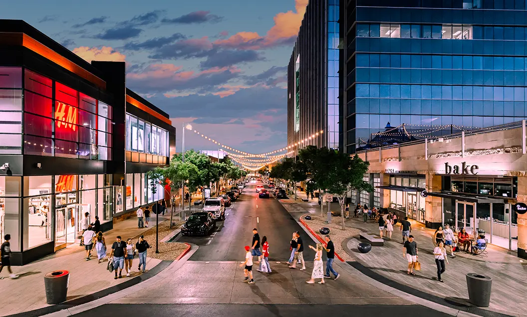 Downtown Summerlin A lively city street at dusk with people walking, shopping, and dining. Storefronts, including H&M and a bakery, line both sides. String lights hang above the street, creating a welcoming atmosphere.