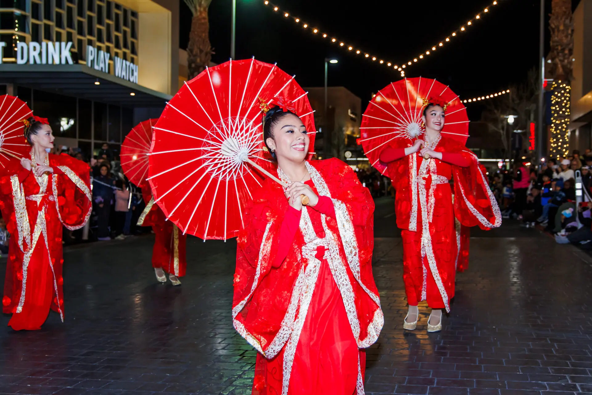 Women in bright red traditional outfits dance with matching red parasols at night during a street festival, with string lights overhead and people watching in the background.