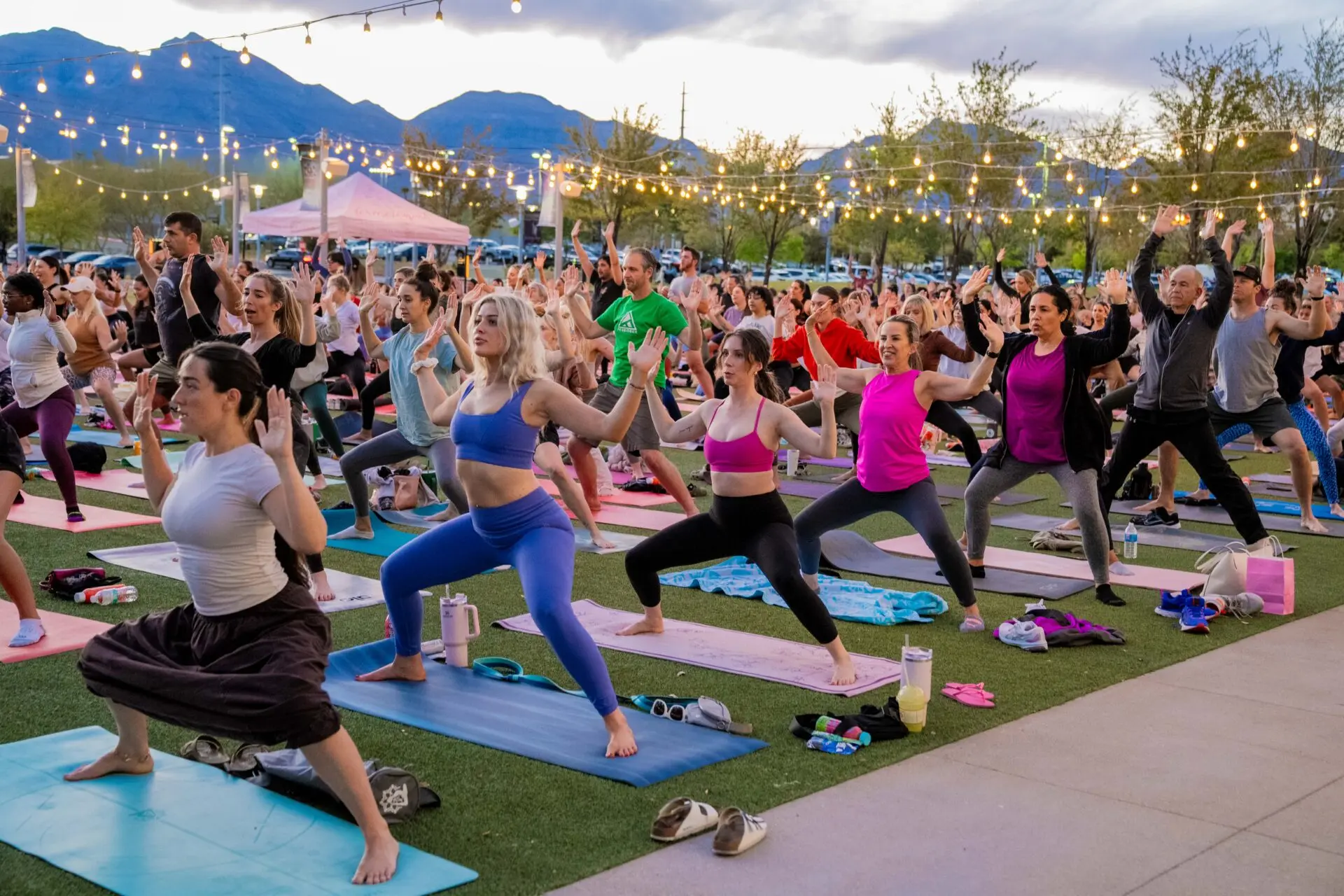 A large group of people practice yoga outdoors on mats at sunset, under string lights, with mountains and trees in the background. Most participants are in a warrior pose, wearing colorful athletic clothing.