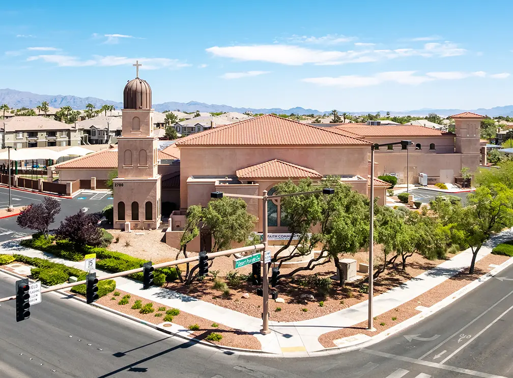 Faith Lutheran Academy Aerial view of a beige stucco church with a bell tower and cross, surrounded by desert landscaping at a city street intersection, with mountains visible in the background under a clear sky.