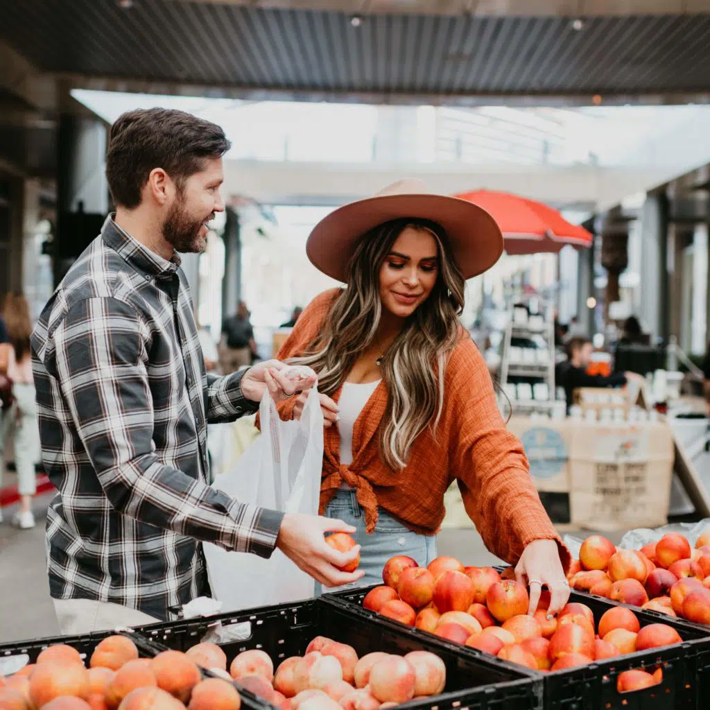 Farmers-Market-at-Downtown-Summerlin-1024×1024 A man and woman select apples from outdoor market crates. The woman wears a wide-brim hat and an orange top, while the man holds a bag and wears a plaid shirt. Other shoppers and stands are visible in the background.