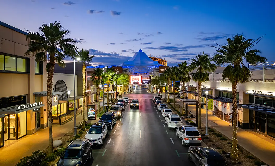 A wide street lined with palm trees, parked cars, and modern shops at dusk, with a large white tent-like structure illuminated in the background.