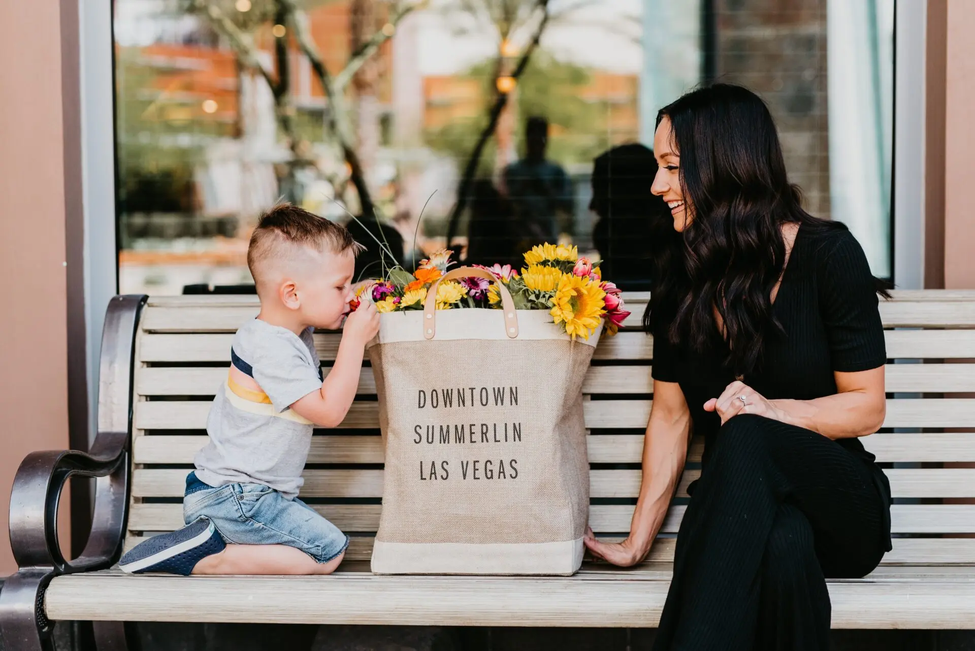 A young boy and a woman sit on a bench with a tote bag filled with flowers that reads Downtown Summerlin Las Vegas. They are smiling and enjoying each others company outdoors.