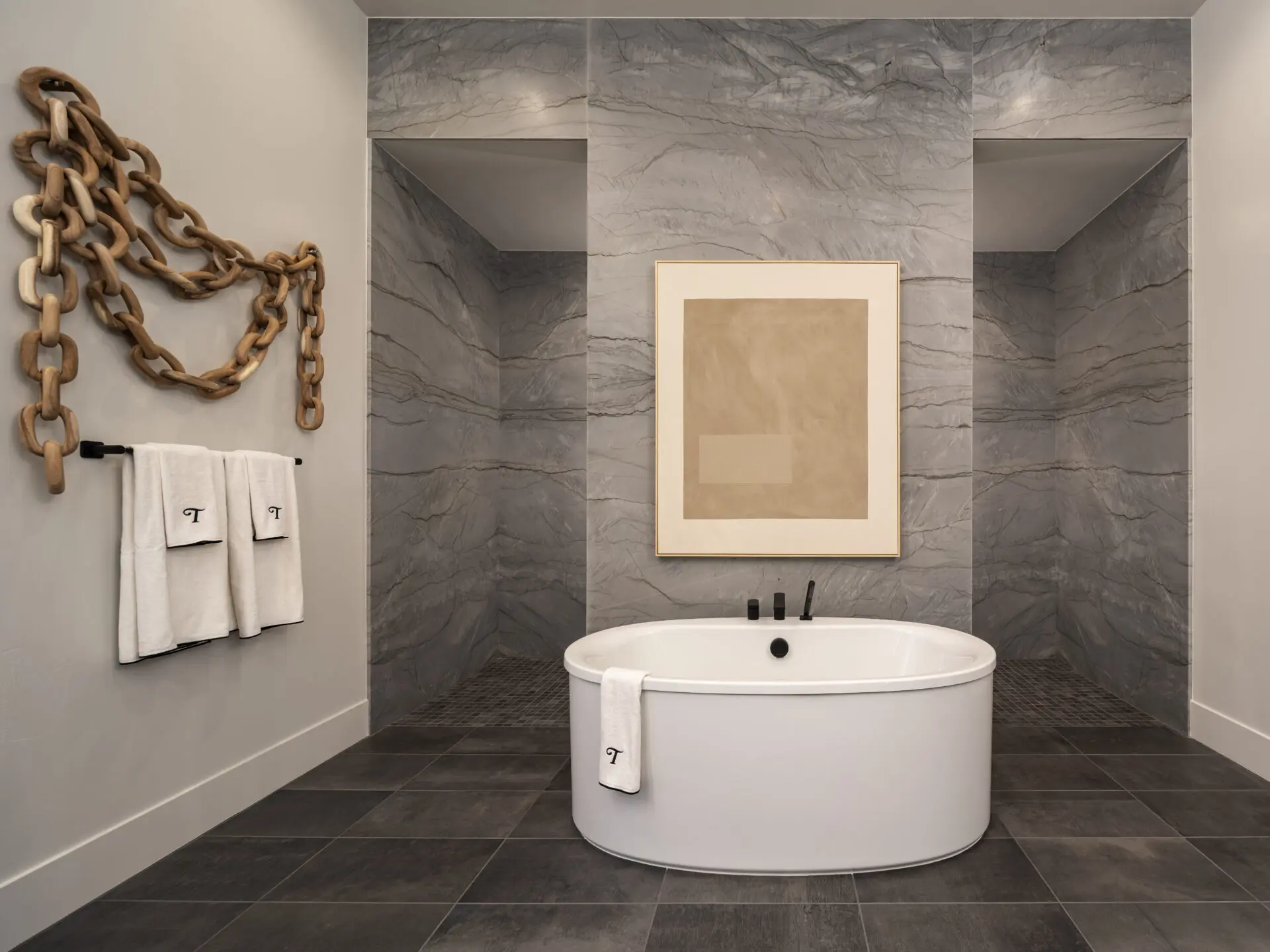 Modern bathroom with a freestanding oval bathtub, gray marble walls, dark tile floor, and minimalist decor including towels, a large abstract art piece, and a decorative wooden chain on the wall.