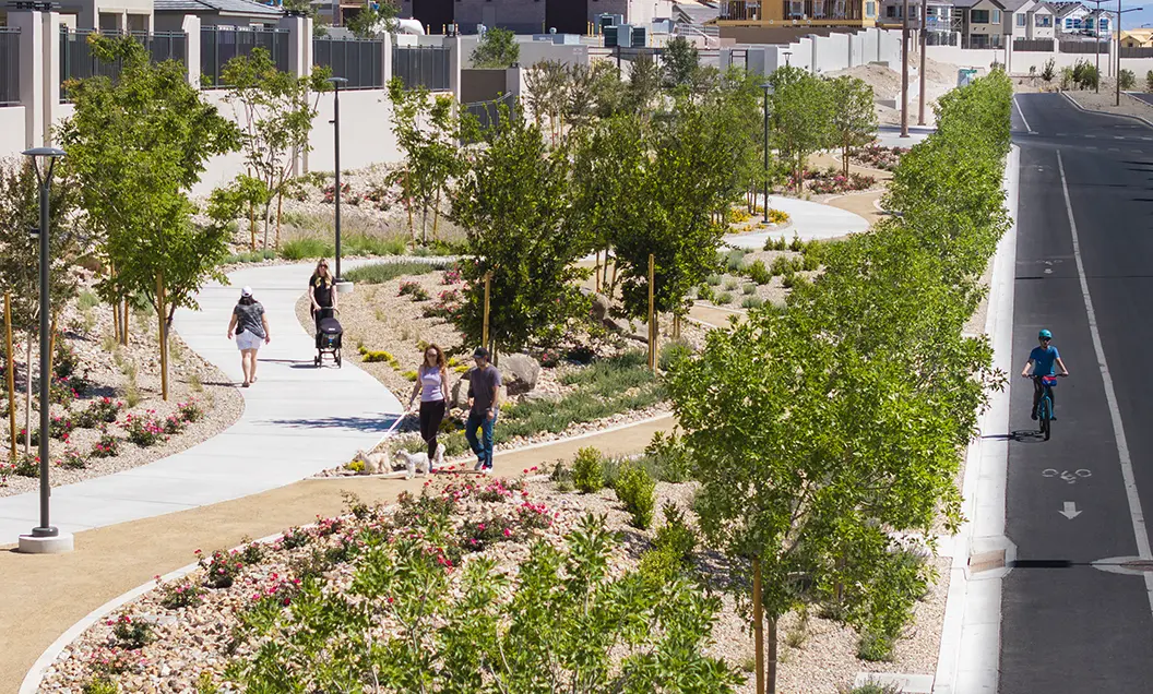 Grand Park Urban Trail 05.2026 – 10 – SELECT A landscaped urban walking path with trees and flowers runs parallel to a street; several people walk, push strollers, and walk dogs on the path, while a cyclist rides in a bike lane on the road.