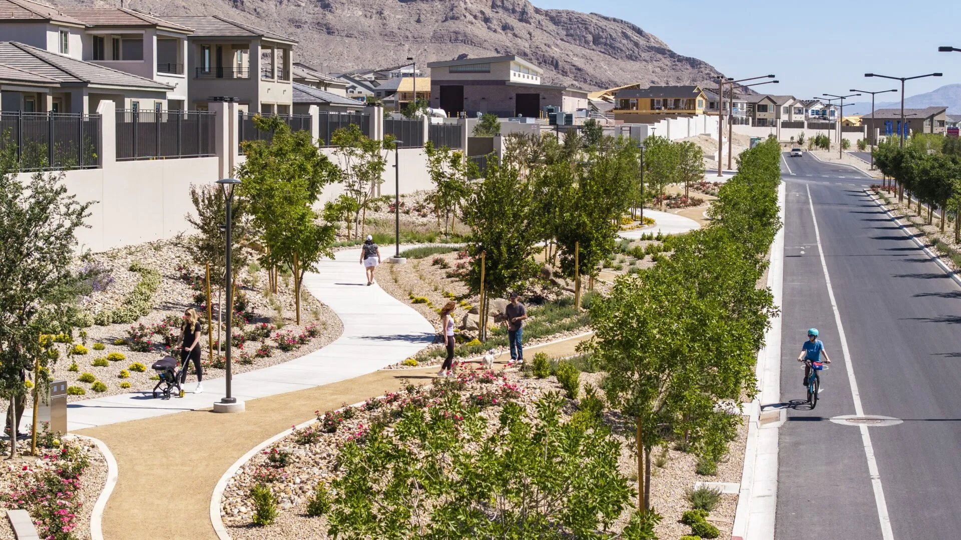 A landscaped suburban street with a winding walking path, trees, flowering plants, and several people walking, jogging, and cycling next to modern houses and mountains in the background.