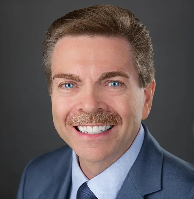 Smiling man with light brown hair, blue eyes, and a mustache, wearing a blue suit, light blue shirt, and navy tie, posed in front of a dark gray background.