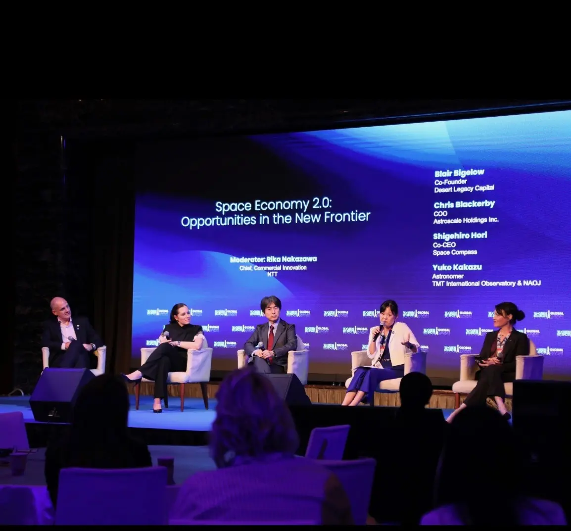 Five panelists sit on stage under a large screen reading “Space Economy 2.0: Opportunities in the New Frontier”; one panelist speaks into a microphone while others listen, with an audience member in the foreground.