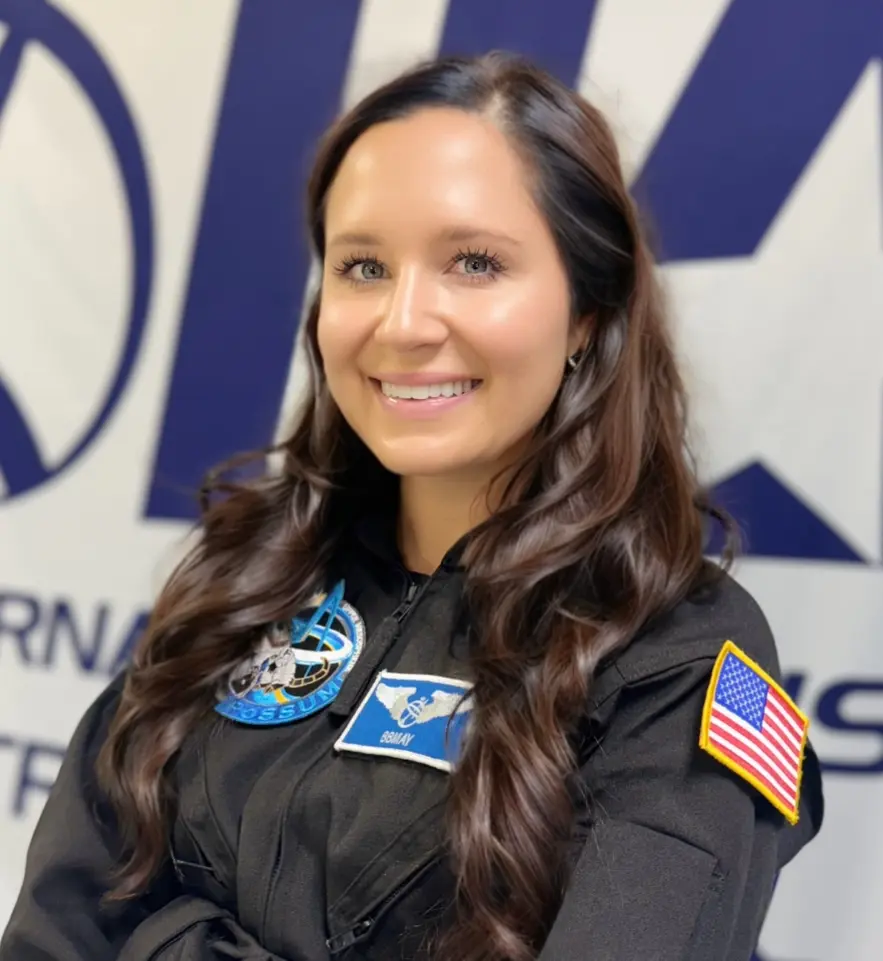 A woman with long brown hair and a black flight suit featuring mission patches and a U.S. flag stands smiling in front of a blurred NASA logo background.