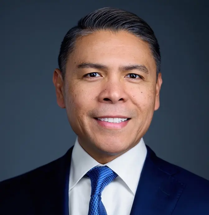 A man in a blue suit, white shirt, and blue tie smiles at the camera against a dark blue background. He has short, neatly combed salt-and-pepper hair.