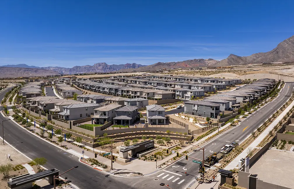 Kestrel Commons Rooftops 05.2026 (5) – SELECT Aerial view of a modern suburban neighborhood with rows of similar gray houses, landscaped yards, and winding streets, set against a backdrop of mountains under a clear blue sky.