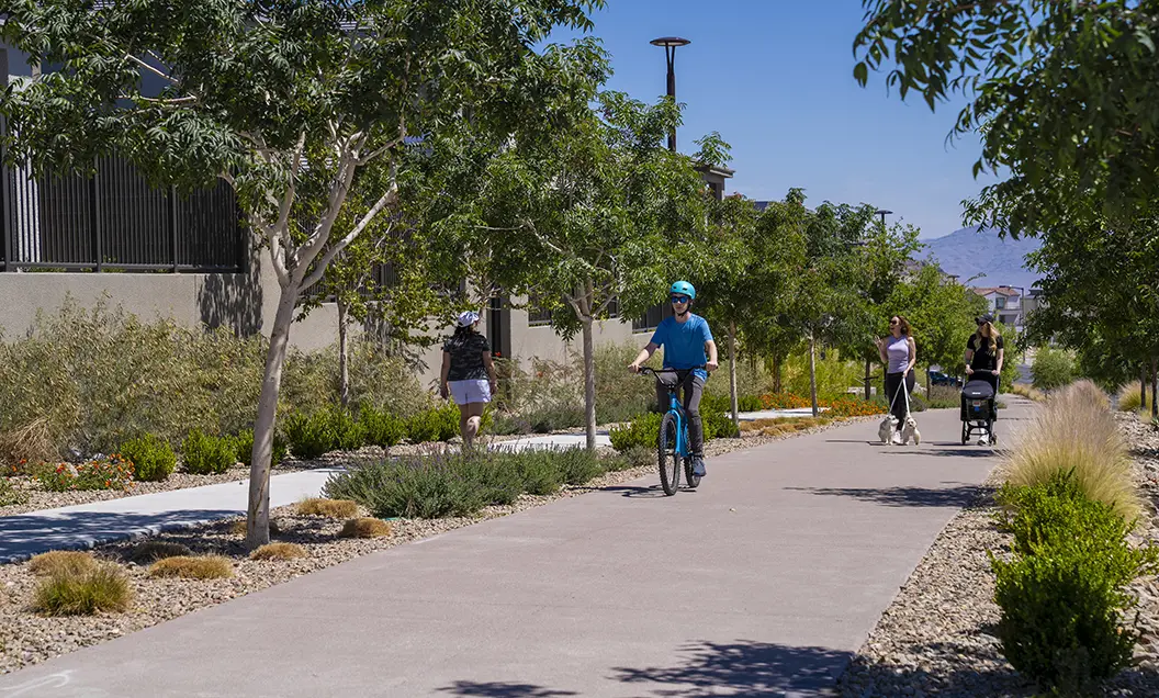 A man rides a bicycle on a paved path lined with trees, while two women walk, one pushing a stroller and the other walking a dog, on a sunny day in a suburban neighborhood.