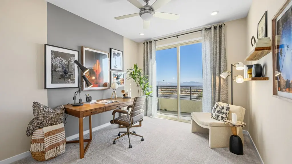 A bright, modern home office features a wooden desk with a chair, wall art, a plant, and a cozy reading nook. Large glass doors let in natural light and reveal a balcony with a scenic outdoor view.