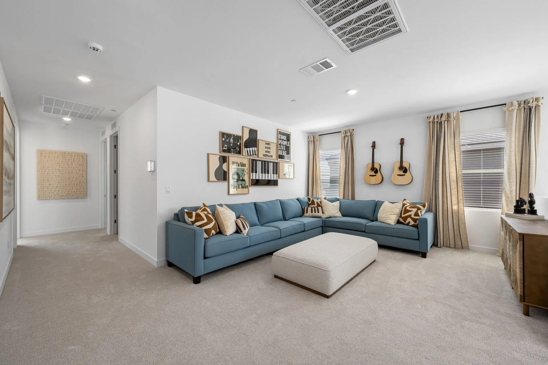A modern living room with a large blue sectional sofa, patterned pillows, a rectangular ottoman, wall art, two acoustic guitars hanging on the wall, beige carpet, and windows with light curtains.